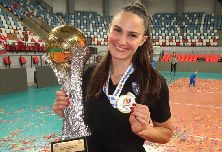 igexportDeorJyugrgh The Volley Lovers Smiling woman in a black shirt holds a tall silver trophy with a gold sphere on top and wears a round medal around her neck, confetti on the floor behind her in an arena.