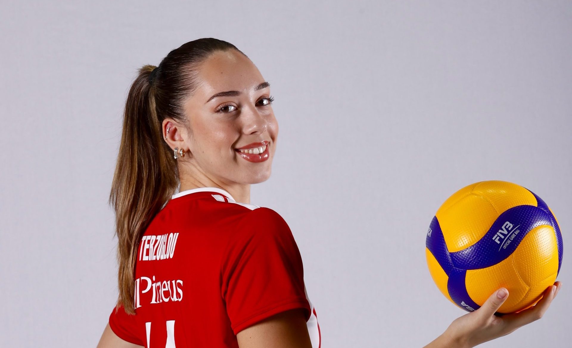 Smiling female volleyball player in a red jersey holding a yellow and blue volleyball.