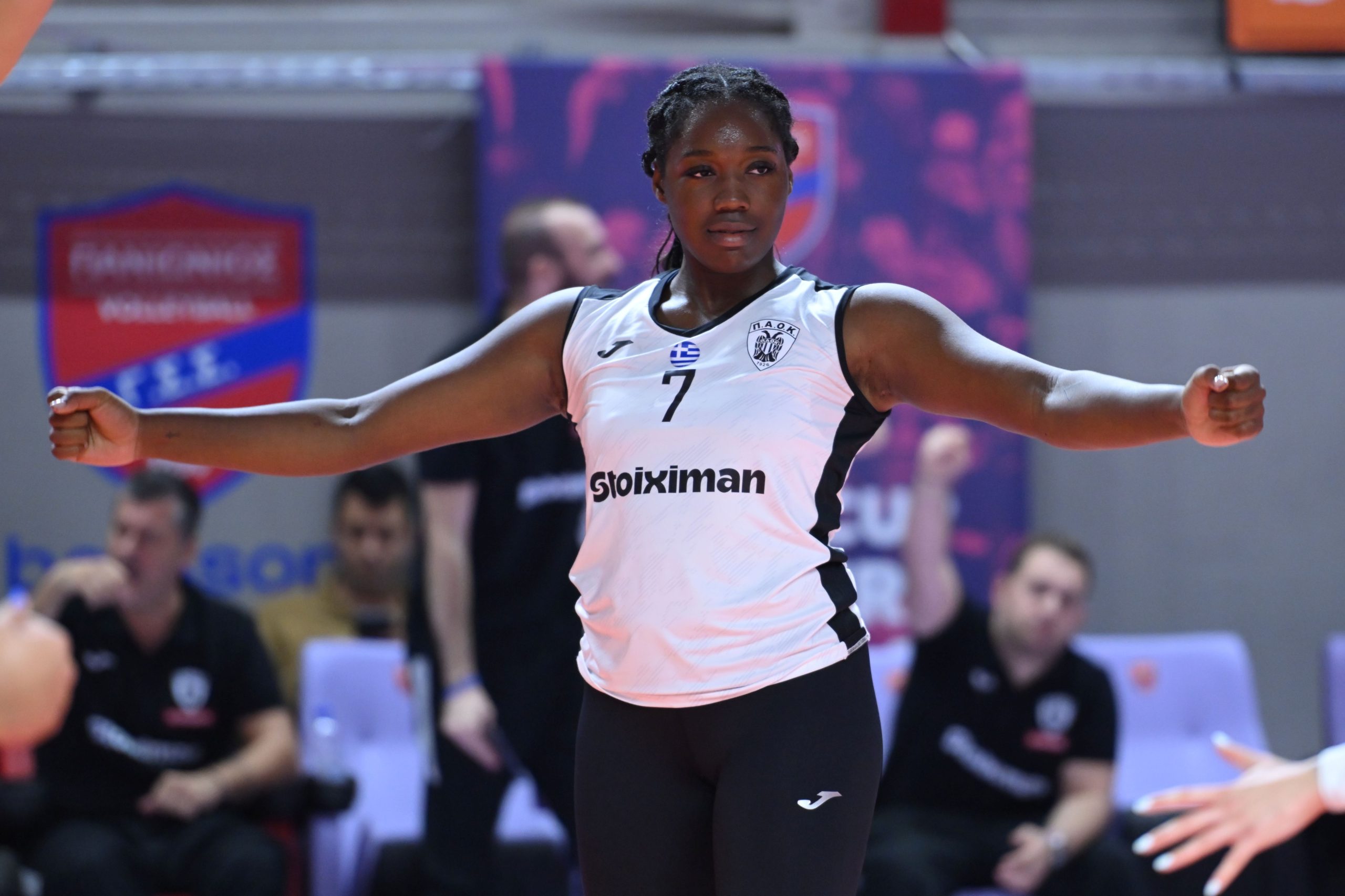 Athlete with arms extended in a match, wearing a white jersey with number 7 and a Stoiximan logo, in an indoor arena with blurred spectators in the background.
