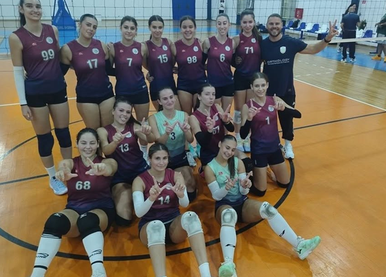 Group photo of a girls' volleyball team posing on an indoor court in maroon uniforms with numbers, some kneeling and others standing, smiling for the camera with a coach nearby.