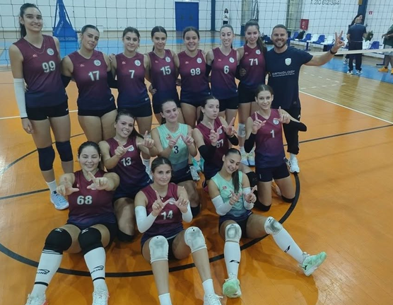 Group photo of a girls' volleyball team posing on an indoor court in maroon uniforms with numbers, some kneeling and others standing, smiling for the camera with a coach nearby.