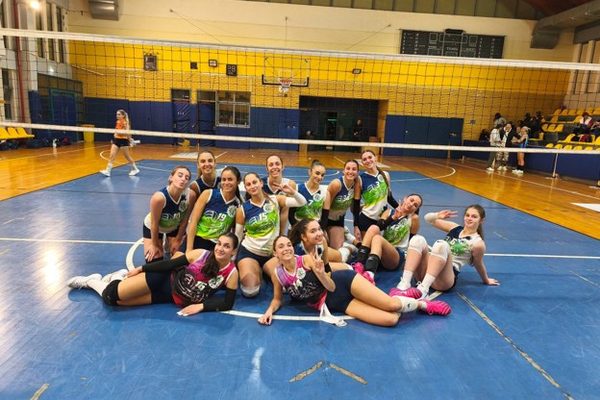 Smiling female volleyball players posing together on an indoor court near the net after a match, wearing matching green and black jerseys.