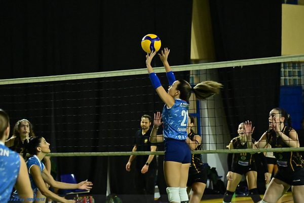 A female volleyball player in blue leaps to spike the ball at the net as teammates and opponents watch nearby.