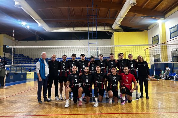 Group photo of a volleyball team posing on the court with coaches, kneeling players in black uniforms with numbers, gym backdrop with blue seating and net behind them.