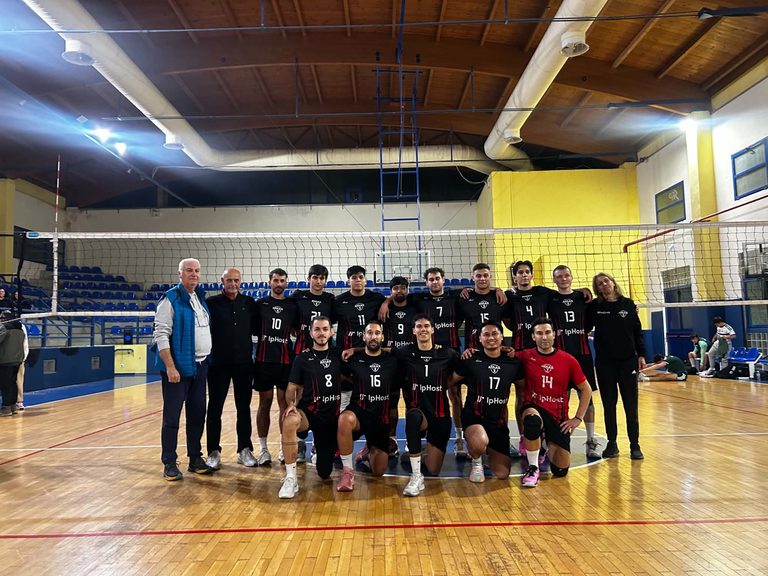 Δελτίο Τύπου 18102026 Β΄ Εθνική Κατηγορία Εθνικό Περιφερειακό Πρωτάθλημα Γυναικών Α΄ Φάση 1η αγωνιστική The Volley Lovers Group photo of a volleyball team posing on the court with coaches, kneeling players in black uniforms with numbers, gym backdrop with blue seating and net behind them.