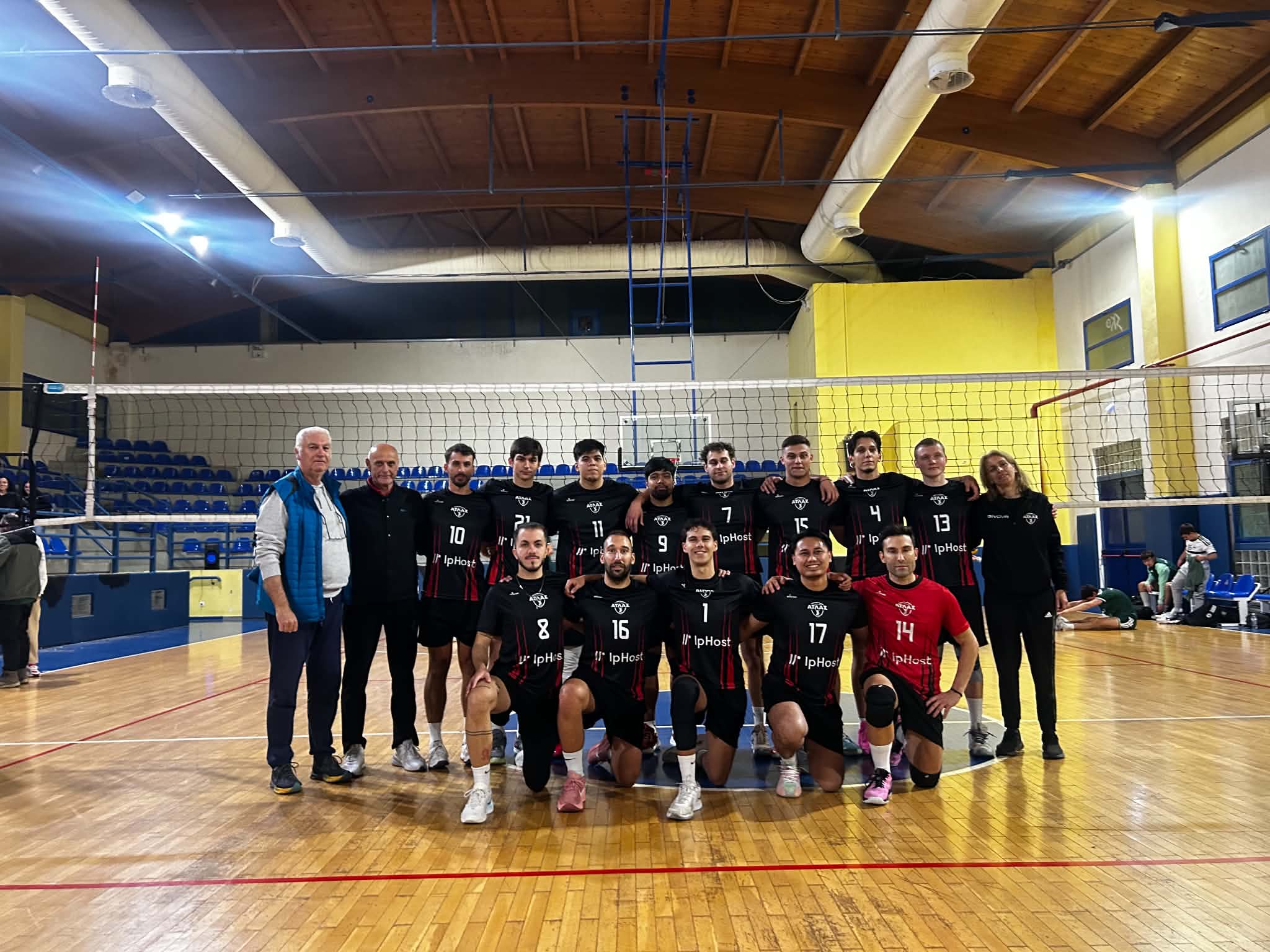 Δελτίο Τύπου 18102026 Β΄ Εθνική Κατηγορία Εθνικό Περιφερειακό Πρωτάθλημα Γυναικών Α΄ Φάση 1η αγωνιστική The Volley Lovers Group photo of a volleyball team posing on the court with coaches, kneeling players in black uniforms with numbers, gym backdrop with blue seating and net behind them.