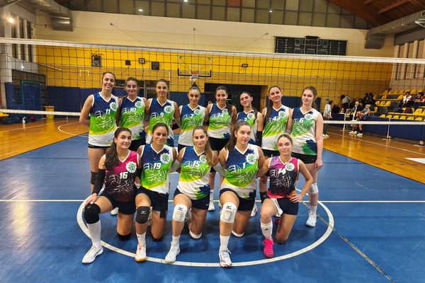 Group of female volleyball players in colorful jerseys posing together on an indoor court with a net in the foreground and bleachers in the background.