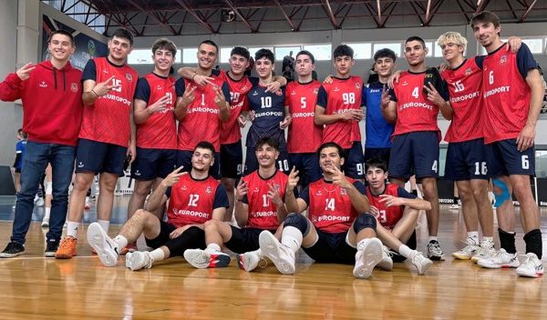 Group of young male athletes in red uniforms posing for a team photo in a gym, two rows (front seated, back standing) with a wooden court floor and arena seating behind them.