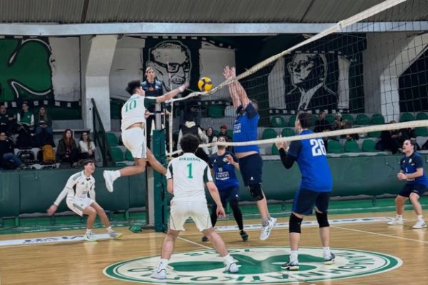 Indoor volleyball match: a white-green team player jumps to spike while blue-team blockers leap at the net; ball suspended in midair near the net.