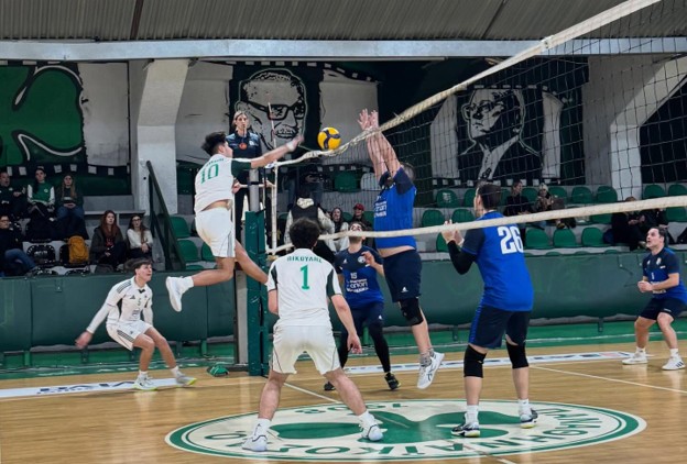 Indoor volleyball match: a white-green team player jumps to spike while blue-team blockers leap at the net; ball suspended in midair near the net.