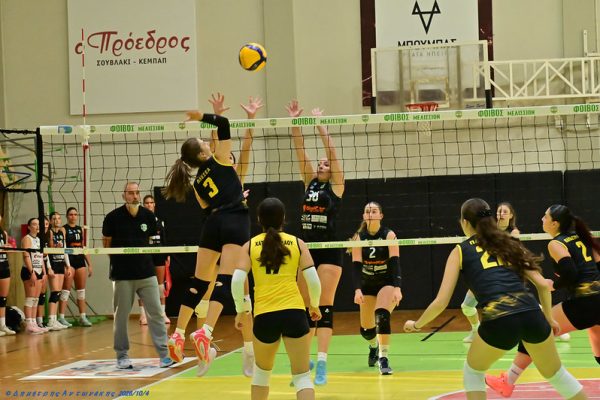 Indoor volleyball match: player in black (#3) spikes the ball over the net as opponents jump to block beneath a green-cushioned court.