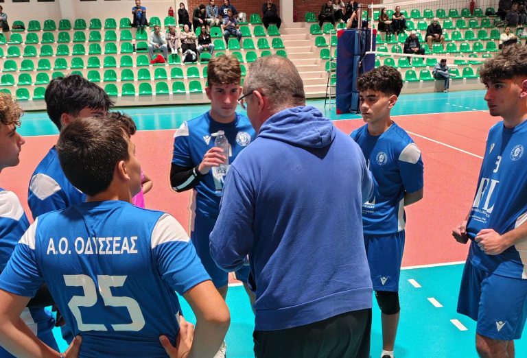 Blue-clad volleyball team huddle around coach on an indoor court, with green empty stands behind.