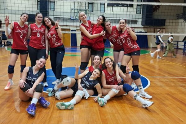 Group of young female volleyball players in red and navy jerseys posing together on an indoor court with a net in the background.