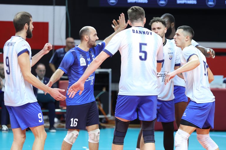 Volleyball players in white and blue uniforms high-fiving in a huddle on a court.