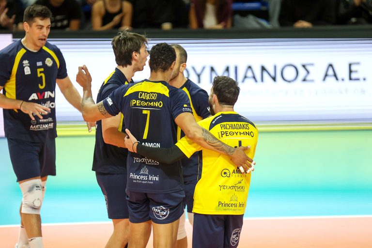 Volleyball players in navy and yellow uniforms celebrate with a group hug on the court.