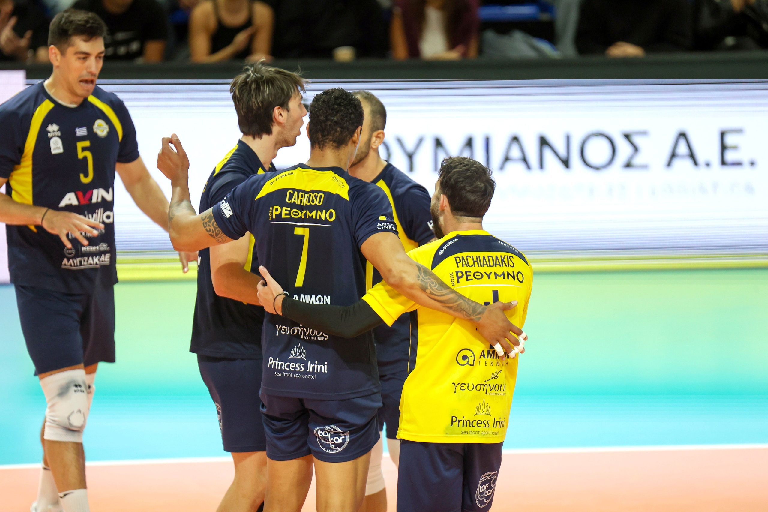 Volleyball players in navy and yellow uniforms celebrate with a group hug on the court.