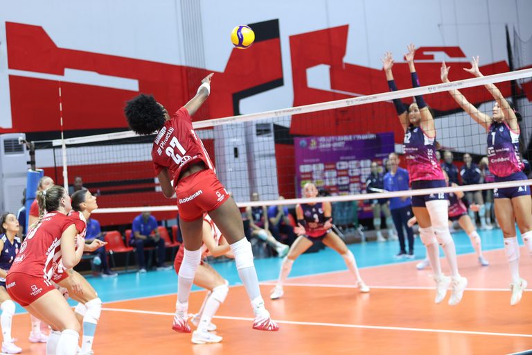 Volleyball player in red jumps to spike the ball over the net as opponents in purple attempt a block indoors.