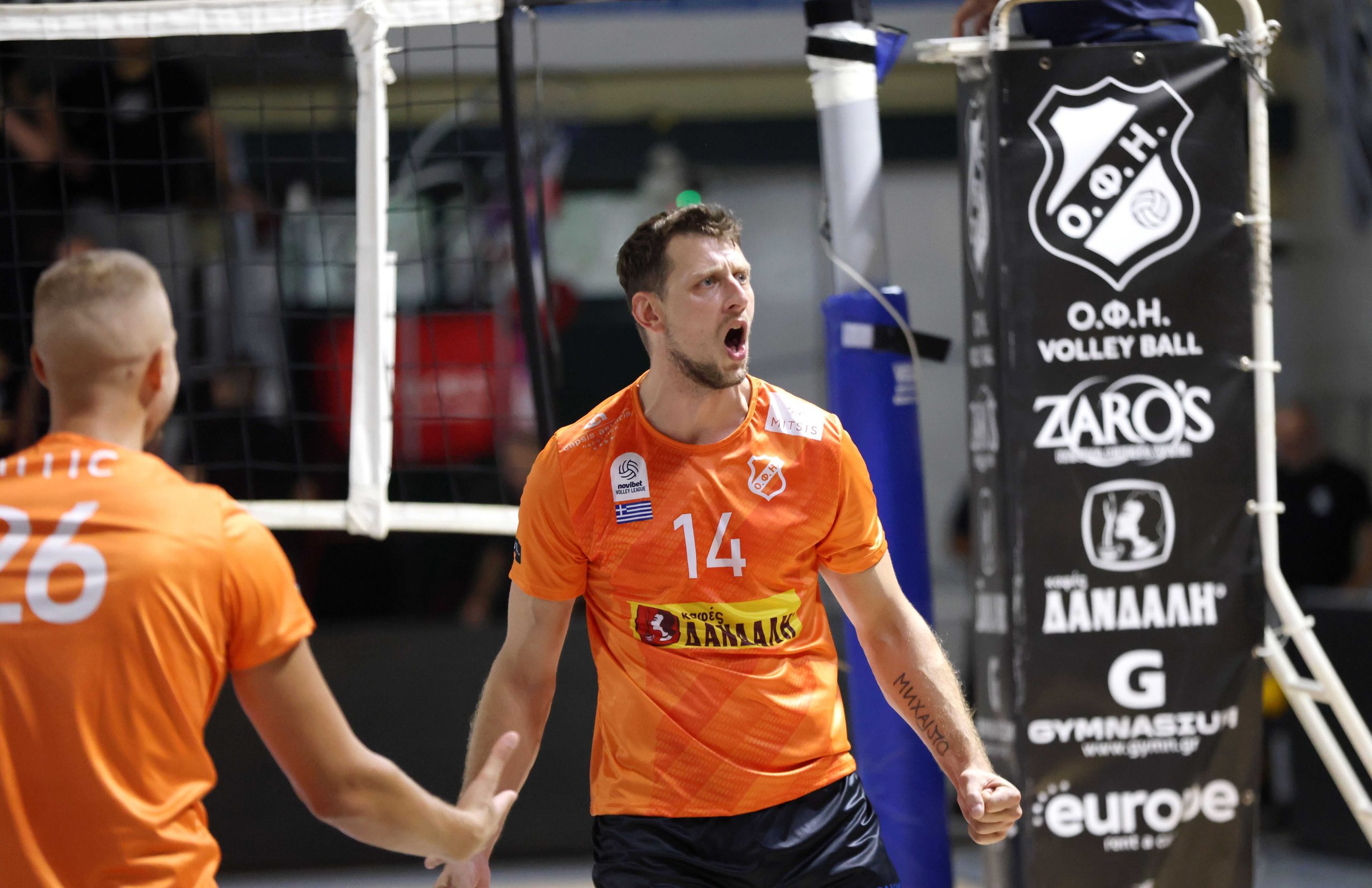 Male volleyball player in orange jersey #14 shouting with mouth open, celebrating a point near the net.