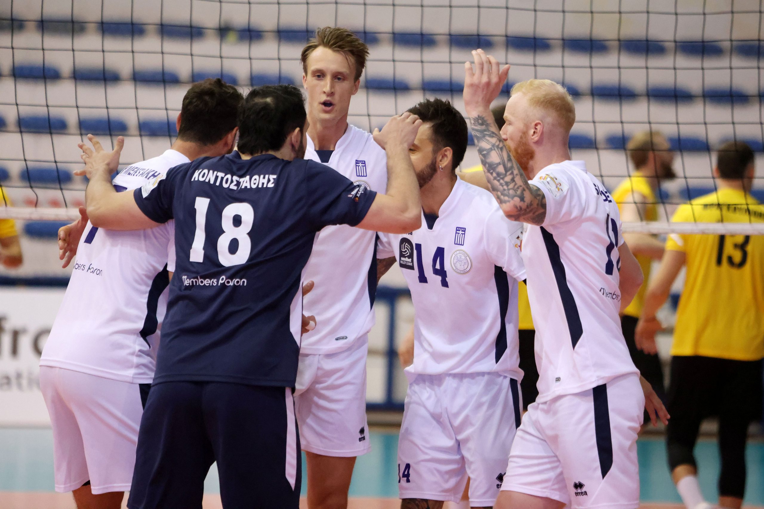 Volleyball players in white and navy uniforms celebrate in a huddle behind the net in a gym.