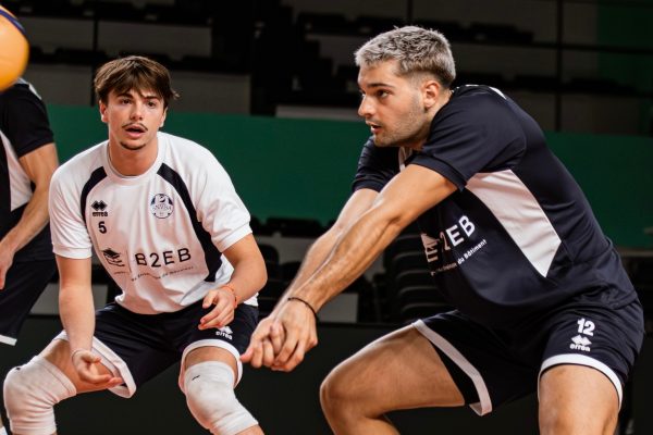 Two male volleyball players in a gymlined scene: one in a white jersey with number 5 and knee braces, the other in a black jersey with number 12, both reaching forward in a defensive stance.
