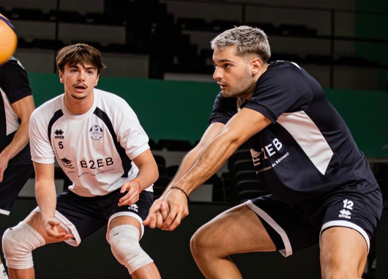 Two male volleyball players in a gymlined scene: one in a white jersey with number 5 and knee braces, the other in a black jersey with number 12, both reaching forward in a defensive stance.